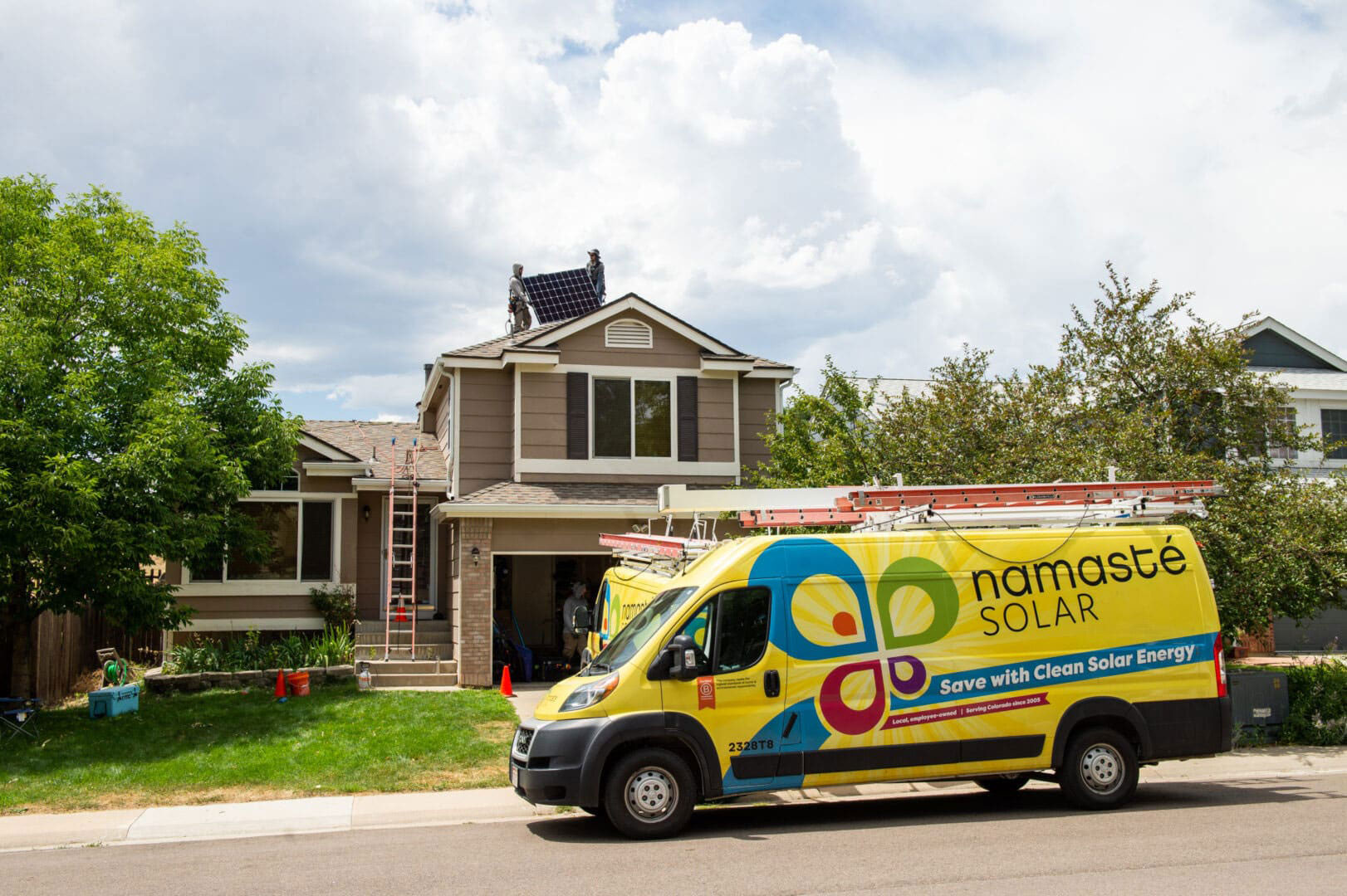 Yellow Namaste Solar van in front of brown house, installers on roof